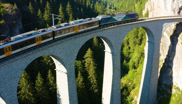 Crossing passenger train traveling over historic stone viaduct through alpine forest gorge