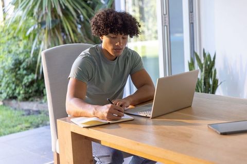 Young man working from home at desk writing in notebook with laptop and indoor plants