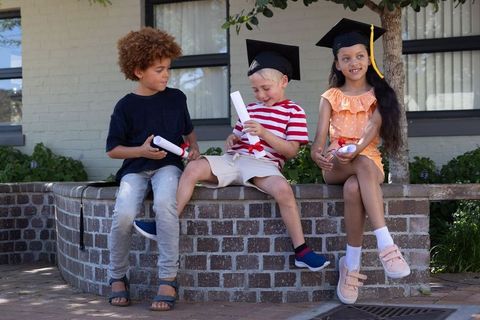 Children celebrating kindergarten graduation sitting on brick courtyard wall with diplomas