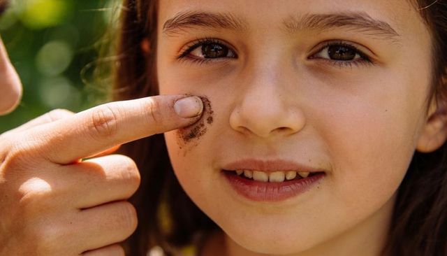 Girl getting dirt on cheek as caregiver finger smearing soil during sunlit backyard play