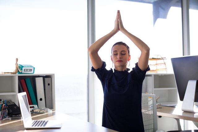 Female executive practicing yoga at her office desk. Perfect for illustrating workplace wellness, stress management, and promoting a healthy work-life balance in corporate environments. Use it in articles, blogs, or presentations about mindfulness, professional well-being, and employee productivity.