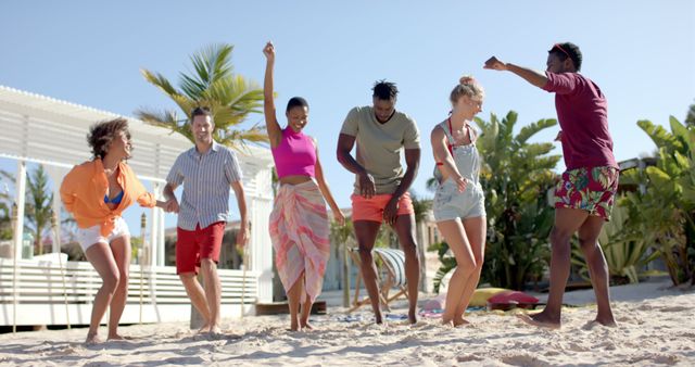 Happy diverse group of friends dancing on beach with beach house and palm trees. Vacation, free time, summer and friendship.