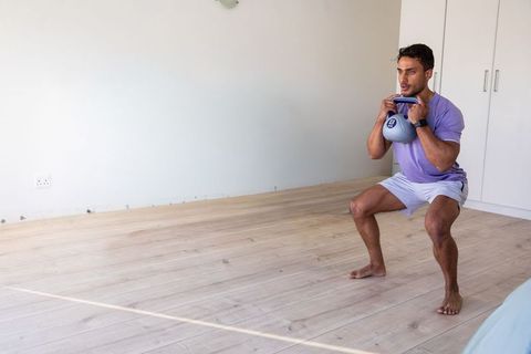 Man performing kettlebell goblet squat in minimalist home workout bedroom, barefoot fitness