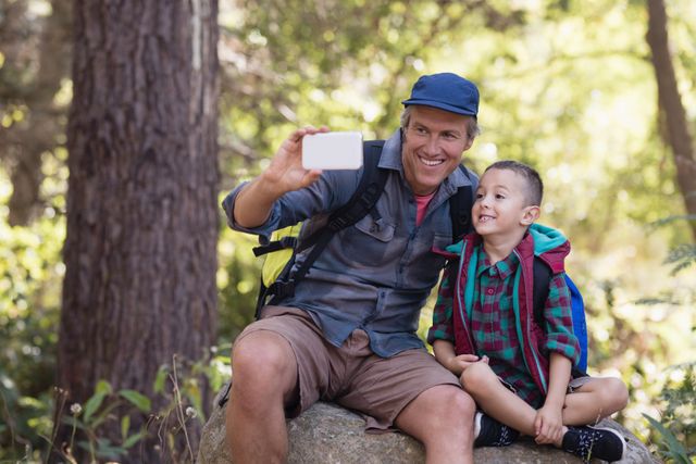 Father and son sitting on rock in forest, taking selfie with mobile phone. Both wearing casual outdoor clothing and backpacks, smiling happily. Ideal for use in family, outdoor adventure, and bonding themes.