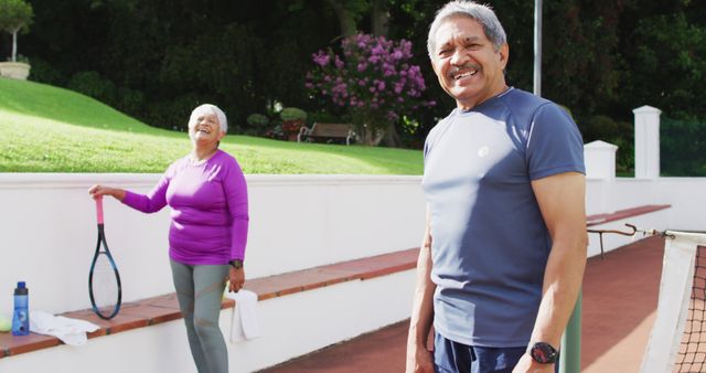 Image of happy biracial senior couple looking at camera on tennis court. active retirement lifestyle, senior relationship and tennis training concept.