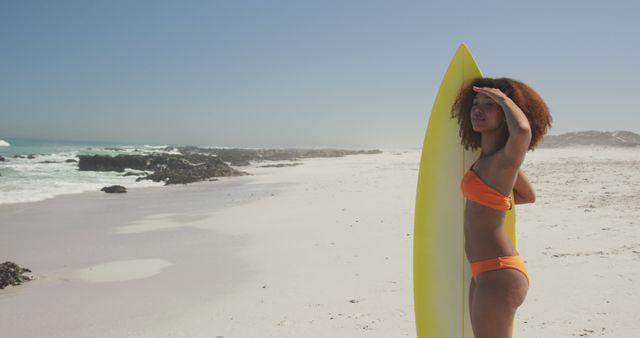 Young woman in orange bikini holding yellow surfboard, looking at ocean. Perfect for promoting beach vacations, surf trips, outdoor lifestyle, summer activities, and beach fashion. Ideal for travel brochures, lifestyle blogs, and summer advertisements.