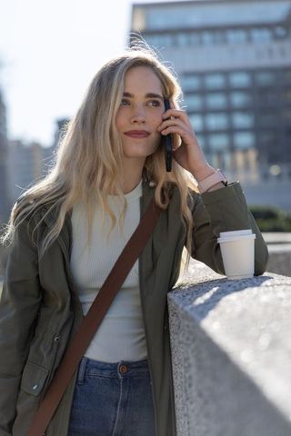Young woman leaning on concrete ledge talking on phone with coffee in urban plaza casual