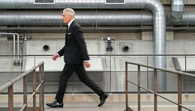 Senior executive striding across industrial walkway in black suit and tie, modern factory interior