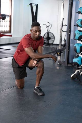 African american athlete kneeling in gym wearing wrist wearable during strength training