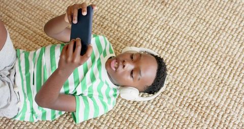 African american boy lying on woven rug listening with headphones and using smartphone