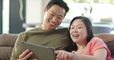 Asian couple relaxing on couch sharing tablet and laughing together in bright living room