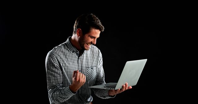 Smiling man in checkered shirt celebrating a successful moment while holding a laptop. Fist pump signifies excitement and victory in a modern, digital setting. Ideal for promotional material related to business success, technology achievements, online learning, and motivational content.
