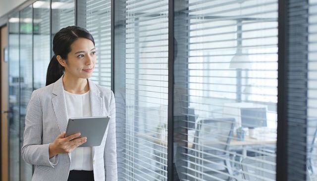 Businesswoman holding tablet and looking through blinds in modern office corridor