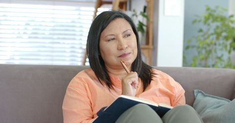 Mature woman sitting on couch journaling and planning, thoughtful cozy home lifestyle