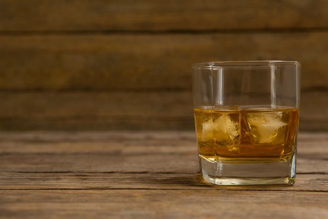 Glass of whisky with ice cubes on a rustic wooden table. Ideal for use in advertisements for alcoholic beverages, bar promotions, or articles about whisky. The warm tones and rustic background create a cozy, inviting atmosphere.