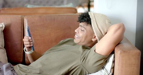 Young man relaxing on leather couch checking smartphone wearing beanie and casual watch