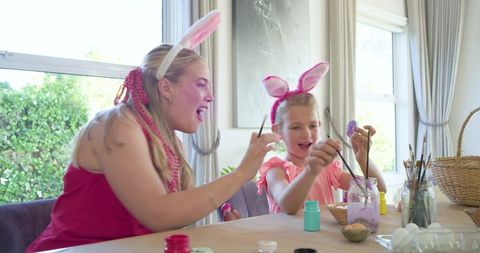 Mother and daughter wearing pink bunny ears painting pastel easter eggs on sunlit table