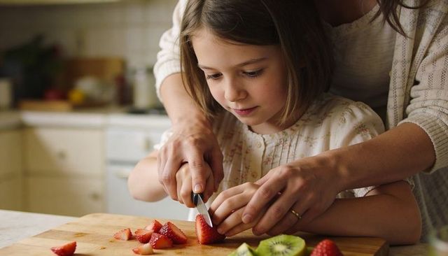 Mother guiding daughter cutting strawberries and kiwi on wooden board home cooking lesson
