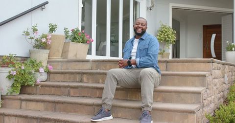 Smiling man sitting on stone steps at home wearing denim jacket and showing wristwatch