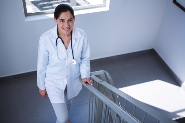 Female doctor walking up a staircase in a hospital, smiling confidently. She is wearing a white coat and has a stethoscope around her neck. This image can be used for healthcare-related content, medical websites, hospital brochures, or articles about medical professionals and their work environment.