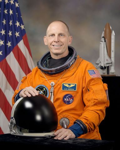 Smiling Astronaut in Orange Spacesuit Holding Helmet Near American Flag and Space Shuttle Model