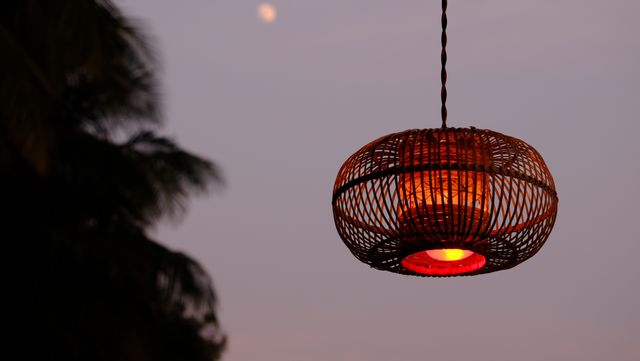Hanging lantern emits a warm glow at dusk with a faint moon and trees silhouetted in the background. This can be used for themes related to tranquil evening settings, outdoor gatherings, atmospheric lighting, or creating a serene ambiance.