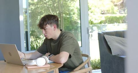 Teen studying on laptop at home dining table with headphones and notebook in daylight