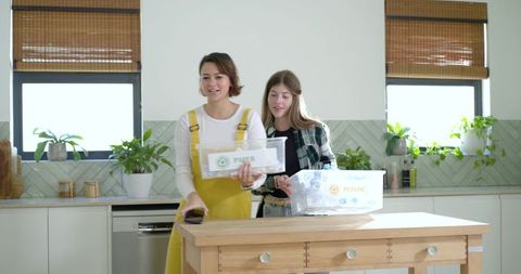 Two women sorting paper and plastic recycling bins on kitchen island, bright lifestyle