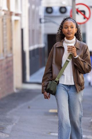 African woman walking on city sidewalk holding takeaway coffee wearing leather jacket