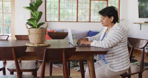 Senior indian woman working from home at dining table typing on laptop with mug and plant