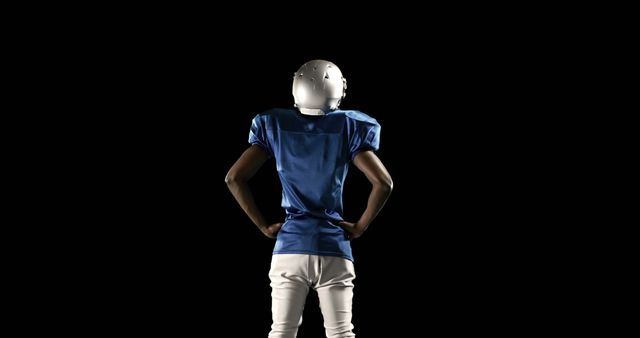 Young male American football player in full gear and helmet stands with hands on hips, back facing the viewer against a dark background. Ideal for sports promotions, athletic gear advertisements, fitness articles, competitive sports blogs, and team spirit campaigns highlighting power, readiness, and determination.