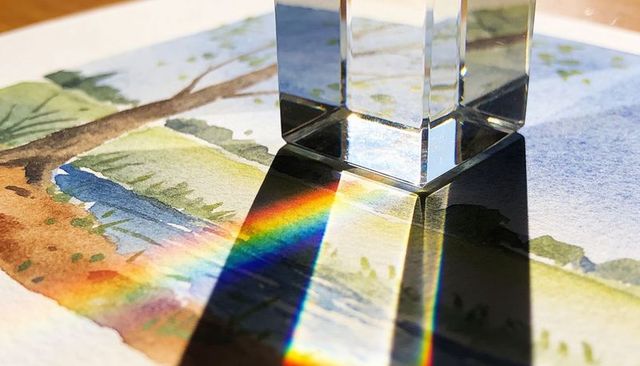 Crystal prism casting rainbow across watercolor tree painting on sunlit table with long shadow