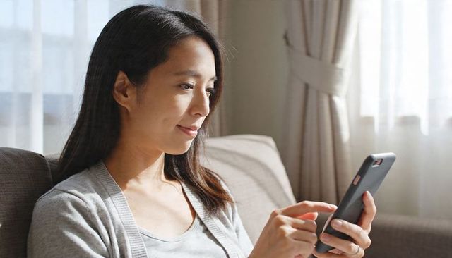 Asian woman holding smartphone checking messages on sofa in soft daylight with curtains