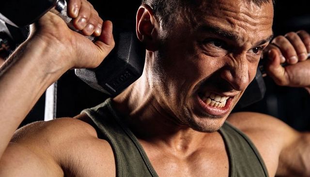 Intense close-up portrait of man lifting heavy dumbbells at shoulders showing strain