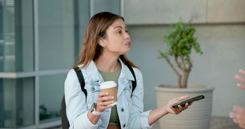 Young woman offering smartphone while holding coffee near modern glass building entrance