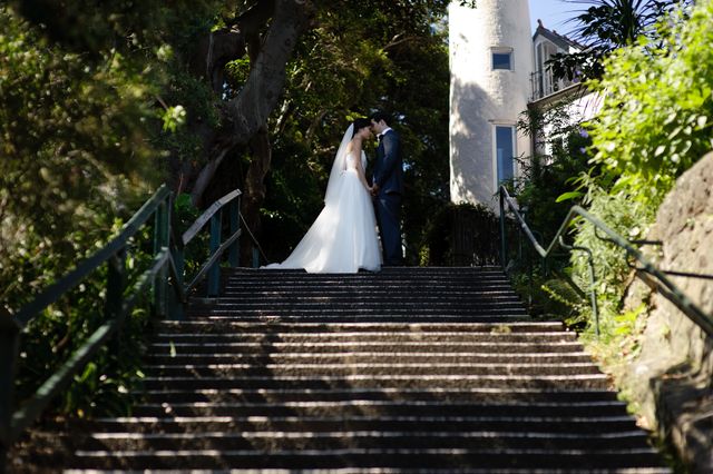 The photo captures a romantic moment of a bride and groom kissing on an outdoor staircase surrounded by greenery. The bride in a white wedding dress and the groom in a suit add an elegant touch to the setting. This image is perfect for use in wedding websites, romantic greeting cards, marriage announcements, or as decoration in a wedding planning blog.