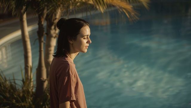 Young woman standing gazing over sunlit pool at golden hour with palm trunks, tranquil