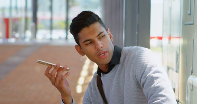 Confident young man casually dressed holding smartphone while in front of modern building. Looks like he is engaging in a conversation or recording a message using his phone. Sunlight and urban environment highlight modern and dynamic lifestyle. Great for use in technology, communication, urban living, and lifestyle concepts.