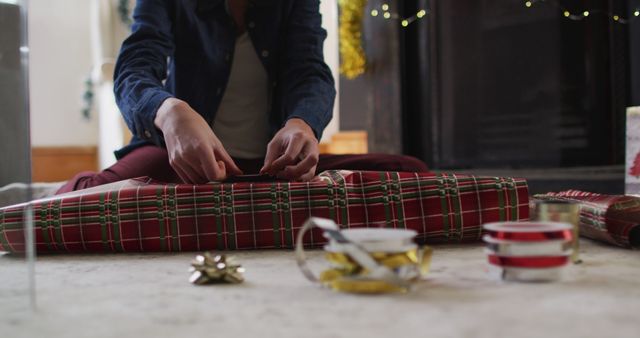 Person sitting on floor wrapping presents with plaid festive paper, with ribbons and bows nearby. Ideal for holiday-themed promotions, DIY gift-wrapping tutorials, festive blogs, and seasonal celebration advertisements.