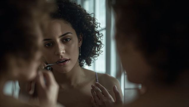 Curly-haired woman brushing teeth in mirror, reflecting in window light, intimate portrait