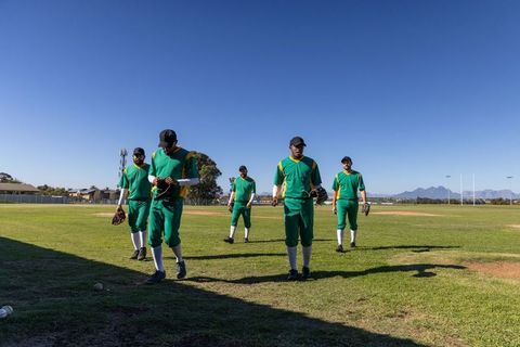 Diverse baseball team walking toward camera on sunny outfield in green uniforms