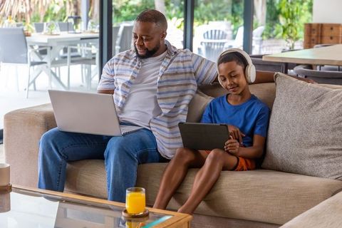 African american father and son using laptop and tablet on living room sofa, bonding