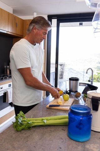 Senior man preparing banana smoothie and chopping fruit in modern minimalist daylight kitchen