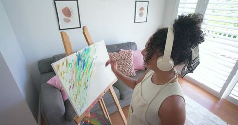 African american woman painting colorful abstract canvas in sunlit home wearing headphones