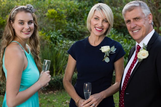 This image shows three smiling wedding guests holding champagne glasses, dressed in formal attire, standing outdoors in a garden setting. Ideal for use in wedding-related content, celebration themes, family gatherings, and event promotions.