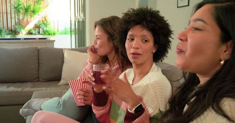 Multicultural women chatting on sofa enjoying popcorn and drinks at cozy hangout in daylight