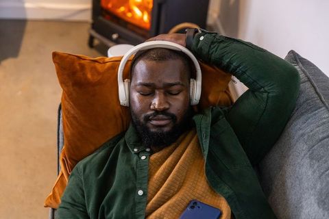 African american man relaxing on sofa with headphones by cozy fireplace at home
