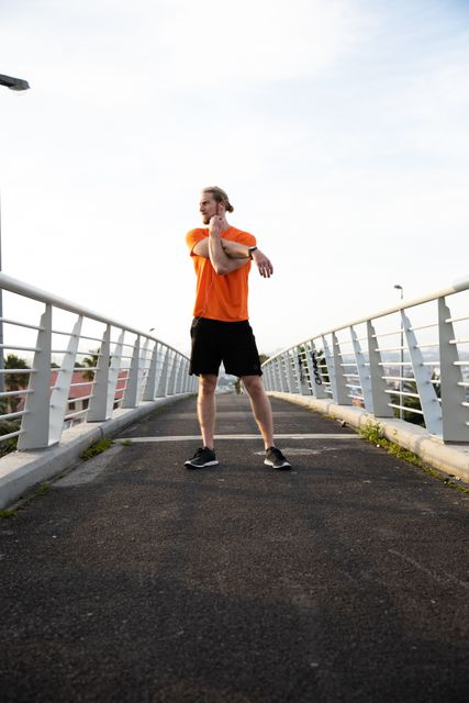 Front view of a fit Caucasian man with long blonde hair wearing sportswear exercising outdoors in the city on a sunny day with blue sky, standing and stretching his arms on a footbridge.
