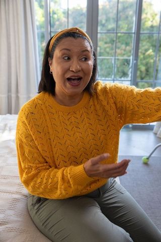 Mature woman wearing mustard sweater talking, gesturing and sitting on bed in bedroom
