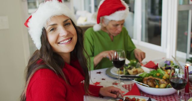 Happy woman wearing Santa hat smiling at camera while enjoying Christmas dinner with elderly family member. Ideal for holiday greeting cards, articles on family celebrations, festive meal advertisements, or emphasizing family togetherness during holiday season.